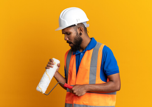 Disappointed young afro american builder man uniform with safety helmet holding looking paint roller isolated orange background with copy space ezgif.com resize (1)